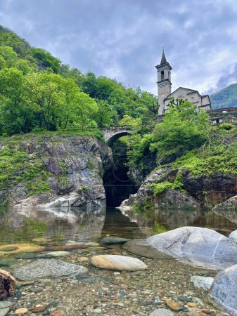 Picturesque view of a historic church with a bell tower beside an ancient stone bridge over a serene river at Orrido di Sant'Anna, surrounded by lush greenery in Traffiume, Cannobio, Piemont, Italy