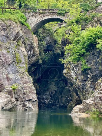 Antike steinerne Bogenbrücke über eine einsame Schlucht mit spiegelndem Wasser, umgeben von schroffen Klippen und üppigem Laub bei Orrido di Sant 'Anna, Traffiume, Cannobio, Piemont, Italien