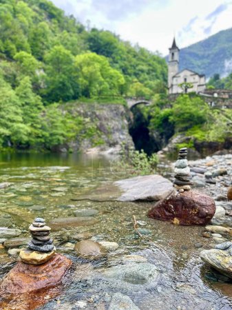 Harmonious blend of nature and heritage: Cairns in a clear mountain stream with Orrido di Sant'Anna church and bridge softly blurred in the background