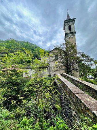 Historische Kirche mit einem hoch aufragenden Glockenturm vor einem dramatisch bewölkten Himmel und üppigem Grün in Orrido di Sant 'Anna, Traffiume, Cannobio, Piemont, Italien