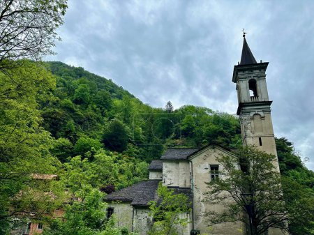 Die majestätische alte Kirche mit dem schlanken Glockenturm schmiegt sich unter wolkenverhangenem Himmel an bewaldete Hügel in Orrido di Sant 'Anna, Traffiume, Cannobio, Piemont, Italien
