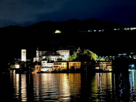Vue nocturne de l'Isola San Giulio avec bâtiments historiques éclairés réfléchissant sur le lac d'Orta, Italie