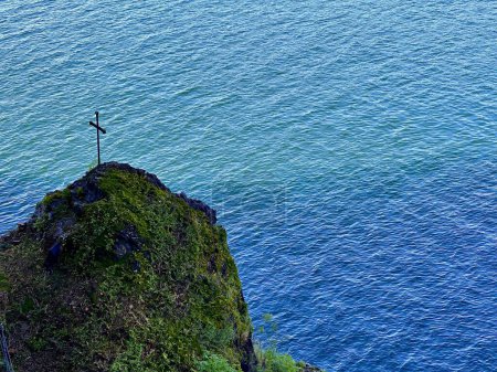 Solitary Cross on Moss-Covered Cliff Overlooking Calm Blue Ocean Waters