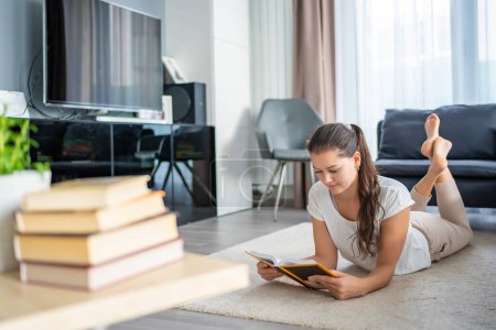 Young woman reads a book lying down and stack of books in the foreground. High quality photo