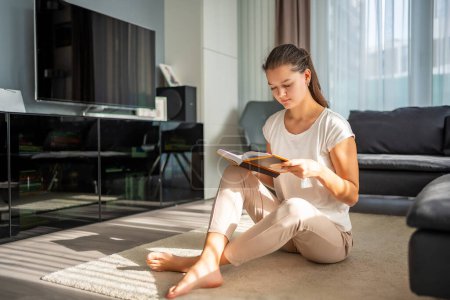 Mujer joven lee libro sentado en el suelo en la sala de estar en el día soleado. Foto de alta calidad