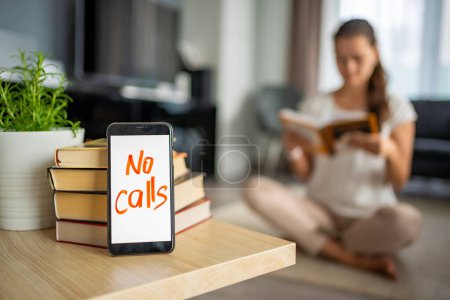 Digital detox concept photo. Smartphone with text No Calls and woman reading book in background. High quality photo