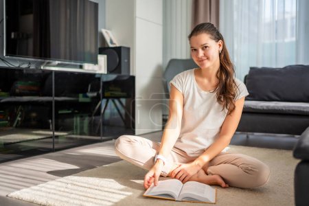 Portrait of smiling young woman reads book sitting on the floor in the living room. High quality photo