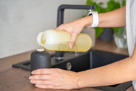 Eine Frau gießt Seife oder Waschmittel aus recycelten Verpackungen in eine Mehrwegflasche in der Küche. Umweltfreundliches Lifestylekonzept. Hochwertiges Foto