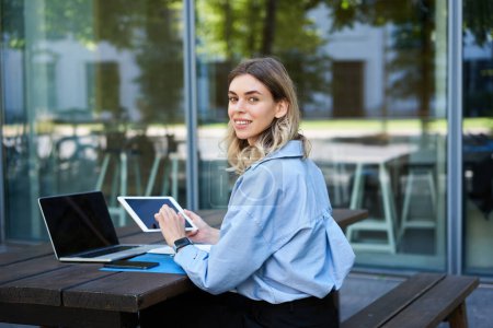 Photo for Portrait of successful businesswoman works outside on fresh air, sits near laptop, holds digital tablet and looks at camera. - Royalty Free Image