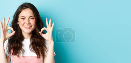 Porträt einer selbstbewusst lächelnden Frau, die vor blauem Hintergrund steht und OK-Geste zeigt, okay sagt, mit Ihnen übereinstimmt, etwas billigt und empfiehlt.