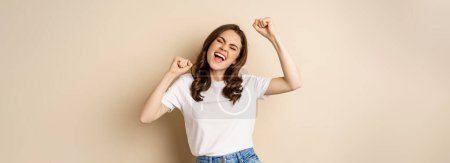 Foto de Entusiasta mujer sorprendida adn ganar, bailar y celebrar, de pie en camiseta sobre fondo beige. - Imagen libre de derechos