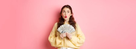 Foto de Pensativo labios pucker mujer joven, mirando a un lado con cara pensativa, sosteniendo billetes de dólar, soñando con comprar algo en dinero, de pie sobre fondo rosa. - Imagen libre de derechos