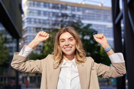Photo for Happy smiling saleswoman in beige suit, celebrates her achievement, triumphing, making fist pump and looking excited on street of business center. - Royalty Free Image