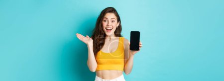 Photo for Attractive young woman in glamour summer clothes, showing empty smatphone screen, gasping amazed, recommending app, standing against blue background. - Royalty Free Image