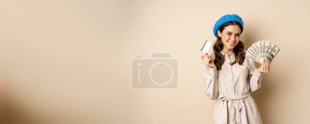 Foto de Microcredit and money concept. Young stylish woman showing credit card and dollars cash, smiling happy and satisfied, standing over beige background. - Imagen libre de derechos