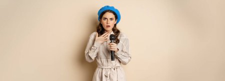 Téléchargez les photos : Image of insecure young woman standing with mic microphone and looking shocked worried, fear of speaking in public, standing over beige background. - en image libre de droit