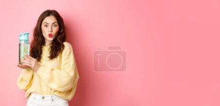 Foto de Joven chica hermosa con el peinado rizado, labios pucker y se ve emocionado, mostrando bebida botella de agua de limón, de pie sobre fondo rosa. - Imagen libre de derechos
