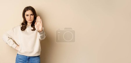 Foto de Stop. Serious and confident woman showing extended arm palm, prohibit, forbid smth, blocking something, standing over beige background. - Imagen libre de derechos