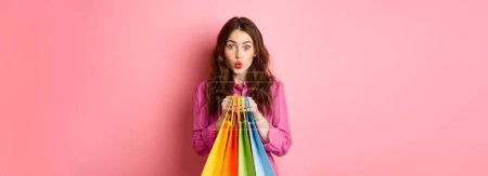 Foto de Glamour chica sosteniendo bolsas de compras, labios pucker y buscando tímido, de pie sobre fondo rosa. - Imagen libre de derechos