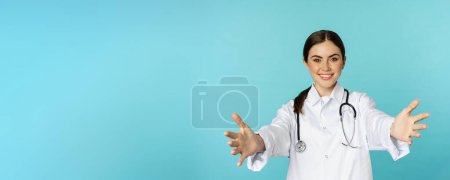 Smiling friendly doctor, girl healthcare worker, intern reaching hands, inviting, hugging or receiving in arms, standing over torquoise background.