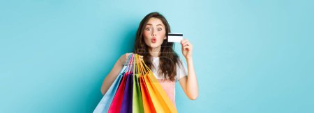 Foto de Chica glamour mostrando tarjetas de crédito de plástico y bolsas de la compra, labios pucker para besar, de pie feliz sobre fondo azul. Copiar espacio - Imagen libre de derechos