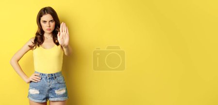 Foto de Serious stylish woman showing stop, rejection, prohibition gesture, decline something, disapprove, standing over yellow background. - Imagen libre de derechos