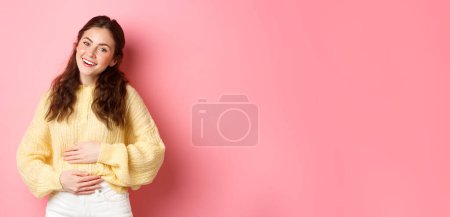 Photo for Young smiling woman touching her stomach with relieved, happy face, feeling good after eating yoghurt or medicine from painful cramps, standing against pink background. - Royalty Free Image