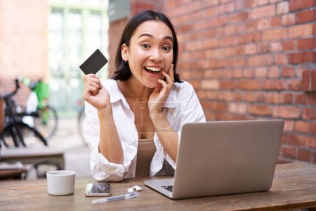 Photo for Portrait of young stylish girl sits near laptop, shows credit card and smiles, pays bills, shops online. shopping and mobile banking concept - Royalty Free Image