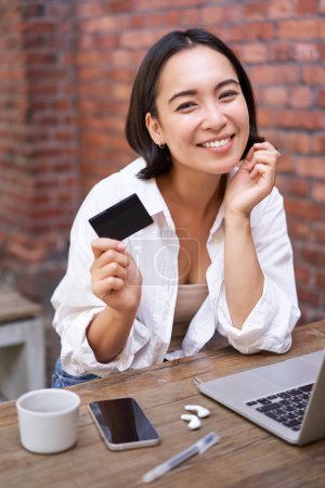 Schöne junge asiatische Frau mit Kreditkarte, sitzt neben Laptop und lächelt, bezahlt Rechnungen, kauft online, bestellt etw. am Computer.