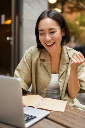 Photo for Vertical shot of happy girl talking on video call, looks at laptop, having online meeting, sitting in outdoor cafe. - Royalty Free Image