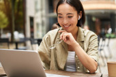 Smiling young korean woman looks at her laptop screen with pleased face, works remotely from outdoors, drinks coffee. Poster #620194986