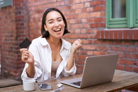 Photo for Enthusiastic asian girl sitting with credit card and laptop, paying contactless, shopping online via computer. - Royalty Free Image
