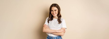 Téléchargez les photos : Portrait of beautiful young woman in white t-shirt, smiling and looking happy, posing against beige background. - en image libre de droit
