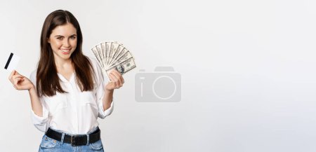 Foto de Happy woman holding money and credit card with thoughtful face, concept of loan and microcredit, standing over white background. - Imagen libre de derechos