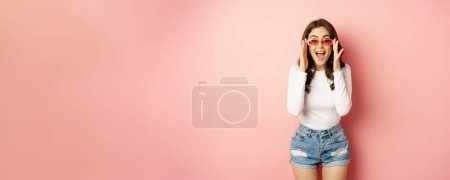 Foto de Enthusiastic brunette girl in stylish sunglasses, reacts surprised and amazed, looking with disbelief at camera, standing over pink background. - Imagen libre de derechos