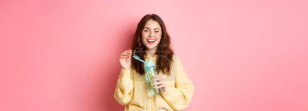 Foto de Retrato de mujer morena feliz con el pelo rizado, beber agua con limón de la botella personal, riendo y sonriendo, sintiéndose saludable, de pie sobre fondo rosa. - Imagen libre de derechos