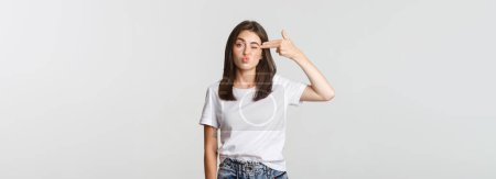 Photo for Unamused cute girl grimacing and showing hand gun gesture at head. - Royalty Free Image