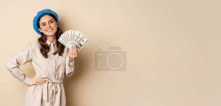 Foto de Stylish young woman holding money, cash dollars, smiling and posing satisfied, going on shopping, standing over beige background. - Imagen libre de derechos