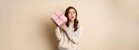 Foto de Excited cute girl shaking box with gift, guessing whats inside present, standing happy against beige background. - Imagen libre de derechos