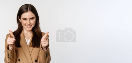 Téléchargez les photos : Enthusiastic businesswoman, corporate woman showing thumbs up and smiling, complimenting, recommending company, standing over white background. - en image libre de droit
