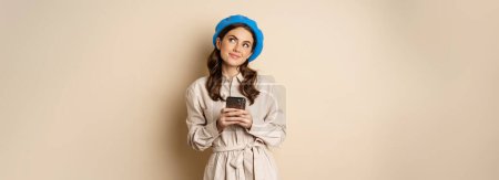 Photo for Dreamy smiling woman in stylish trenchcoat, looking up fantasizing while shopping on mobile phone app, using smartphone and thinking, standing over beige background. - Royalty Free Image