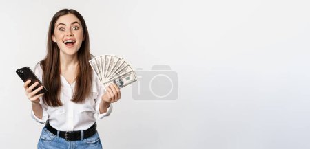 Foto de Online microcredit loans and banking concept. Happy woman holding mobile phone and money, smiling and laughing, standing over white background. - Imagen libre de derechos