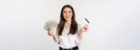 Foto de Happy woman holding money and credit card with thoughtful face, concept of loan and microcredit, standing over white background. - Imagen libre de derechos