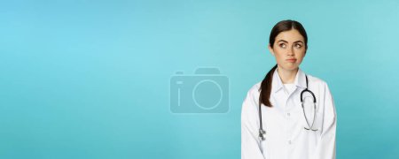 Photo for Image of woman doctor, female medial staff in white lab coat, looking away thoughful, making decision, thinking of smth, standing over blue background. - Royalty Free Image