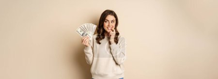 Foto de Excited brunette woman holding money and biting finger with yearning face, watns to buy smth, shopoholic with cash, standing over beige background. - Imagen libre de derechos