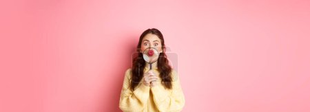 Foto de Mujer tonta coqueta mostrando sus labios pucker, besándose la cara con lupa cerca de la boca, mirando divertido a la cámara, de pie sobre el fondo rosa. - Imagen libre de derechos