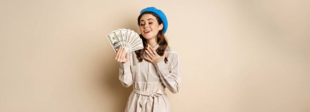 Foto de Stylish young woman holding money, cash dollars, smiling and posing satisfied, going on shopping, standing over beige background. - Imagen libre de derechos