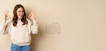 Photo for Very good, nice work. Cheerful young woman agrees, shows okay, ok praise, recommending smth, standing over beige background. - Royalty Free Image
