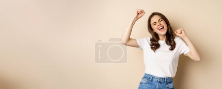 Foto de Entusiasta mujer sorprendida adn ganar, bailar y celebrar, de pie en camiseta sobre fondo beige. - Imagen libre de derechos