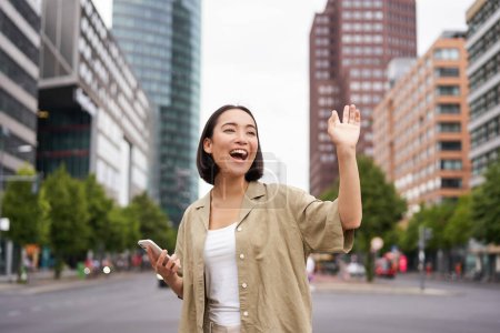 Téléchargez les photos : Heureuse fille asiatique passant par un ami et les saluant dans la rue, disant bonjour tout en marchant dans la ville, tenant smartphone. - en image libre de droit